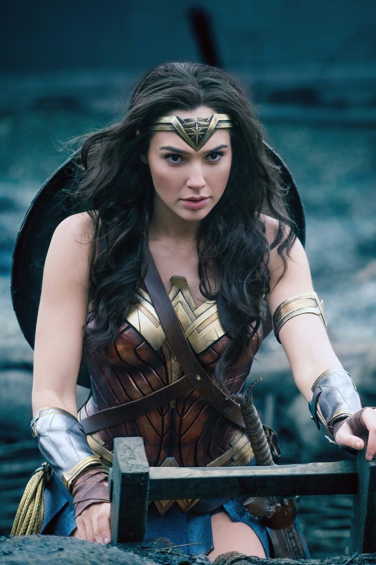 this image features wonder woman, played by gal gadot, in her iconic costume from the 2017 movie wonder woman. she is standing in a trench with a shield on her back and a sword at her side. she is looking intently at something offscreen, her face determined and focused. the image captures the essence of wonder woman as a powerful and inspiring superhero.