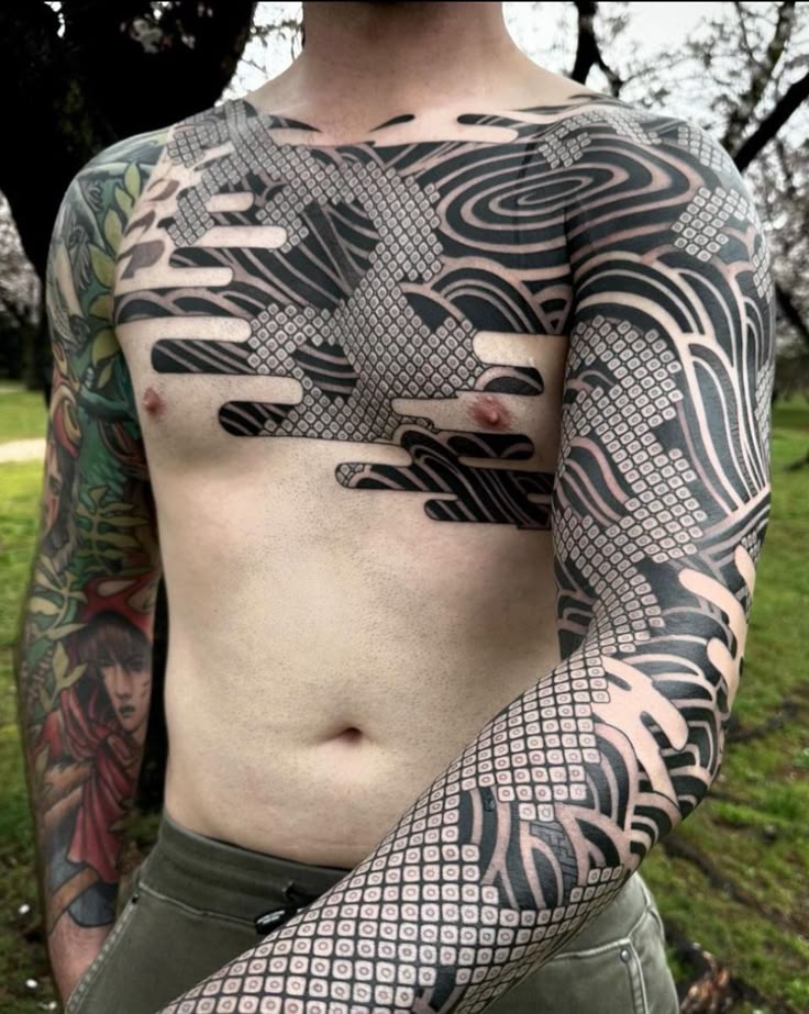 a shirtless man with extensive black ink tattoos covering his chest, arms, and torso. the tattoos feature traditional japanese designs with water patterns and grid patterns. one arm also has a tattoo with a redrobed figure. he is standing outdoors in a grassy area with trees in the background.