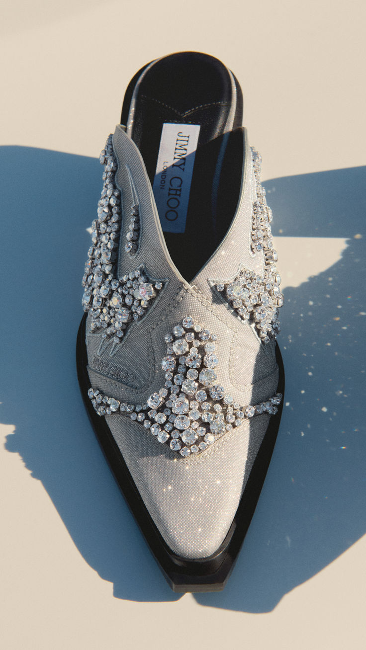 a closeup, highangle shot of a jimmy choo mule. the shoe is a gray, cowboyinspired design and is heavily embellished with crystals. the black insole shows the jimmy choo logo. the shoe is sitting on a lightcolored surface, casting a shadow to the left.