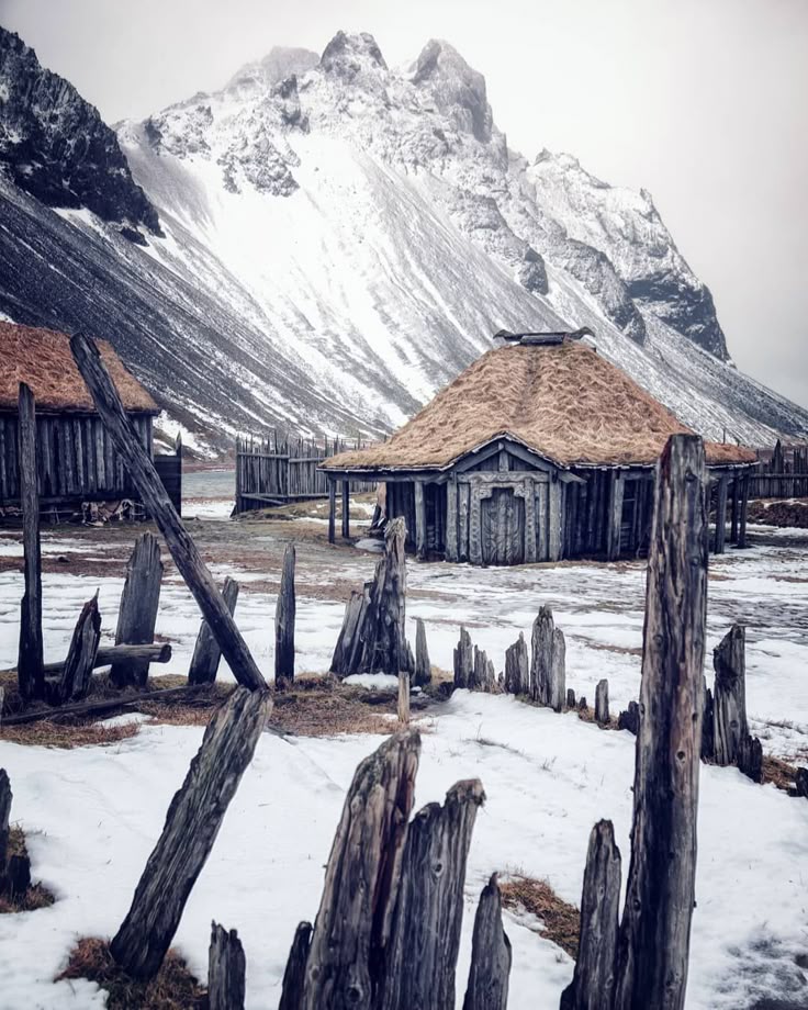 the image shows a village scene with thatchedroof huts set against a backdrop of snowcovered mountains. the scene takes place in the winter, with snow visible on the ground and surrounding structures. wooden fences and posts surround the huts. the sky is overcast, and the overall mood is cold and desolate. the location may be iceland.