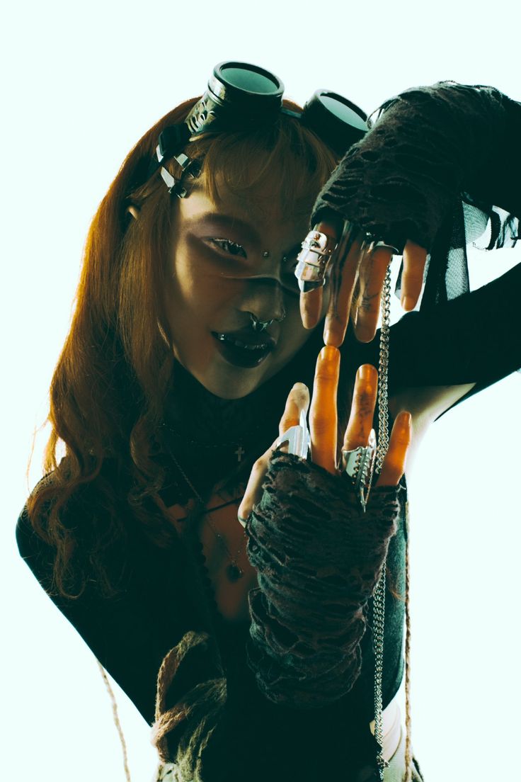 a woman with red hair is wearing dark clothing and dark goggles. she is looking at the camera and her hand is in front of her face. the image is dark and moody.  the woman is wearing numerous accessories.