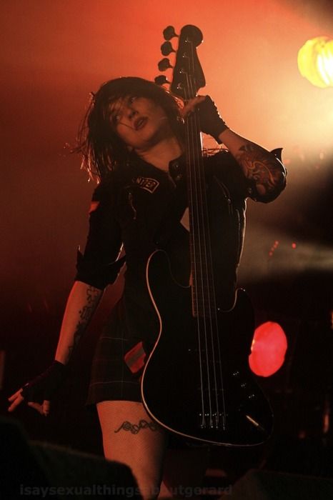 a closeup shot of a female musician passionately playing a bass guitar on a dimly lit stage during a concert. she is wearing a black leather jacket, dark shorts, and has tattoos visible on her arms and legs. the atmosphere is intense and energetic. the red lighting adds to the dramatic mood.