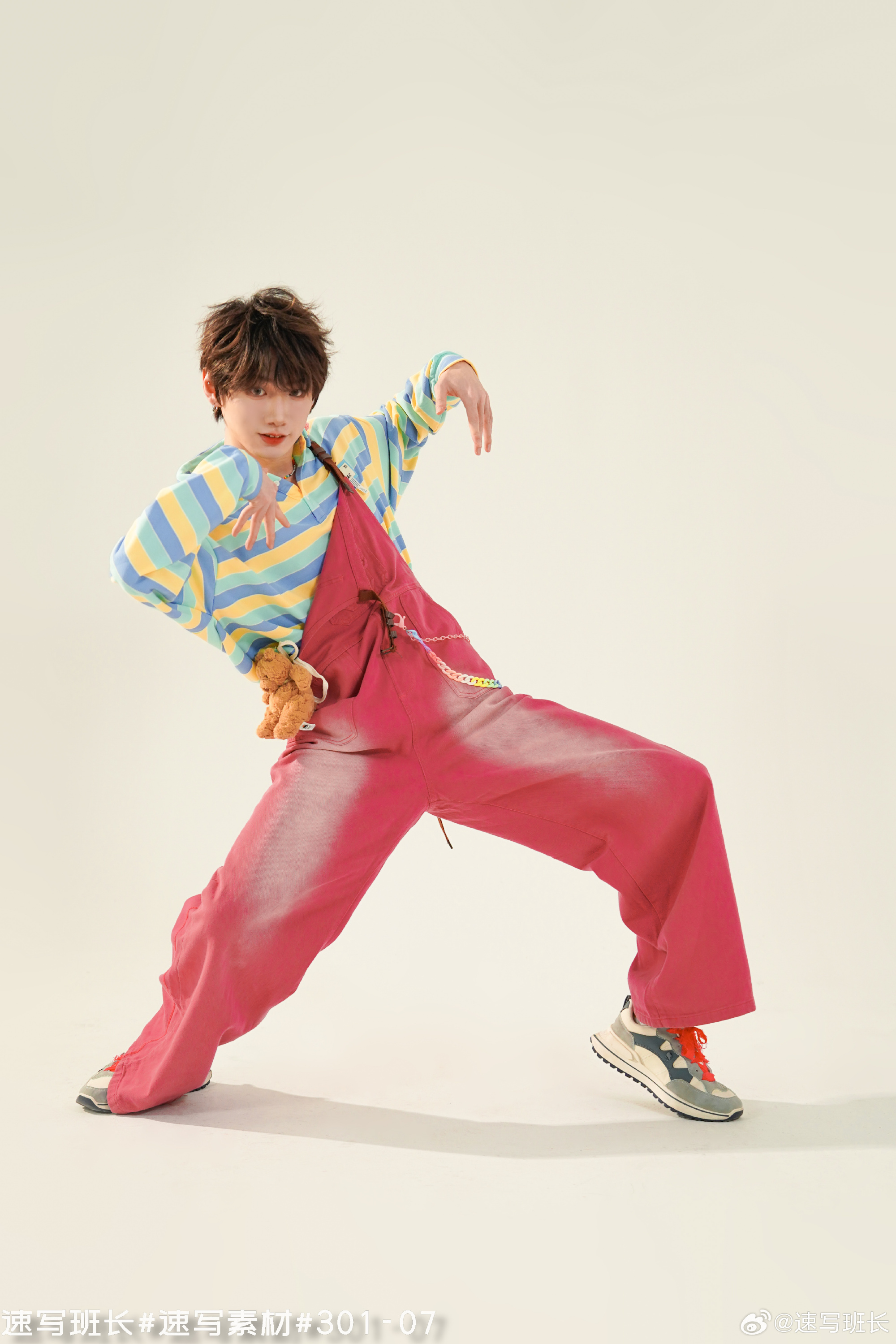 a young asian man in a playful, colorful outfit is captured middance pose against a white studio background. he is wearing a striped, pastelcolored sweatshirt, bright pink overalls, and sneakers. the bright, artificial lighting illuminates the scene and emphasizes the details of his clothing and pose.