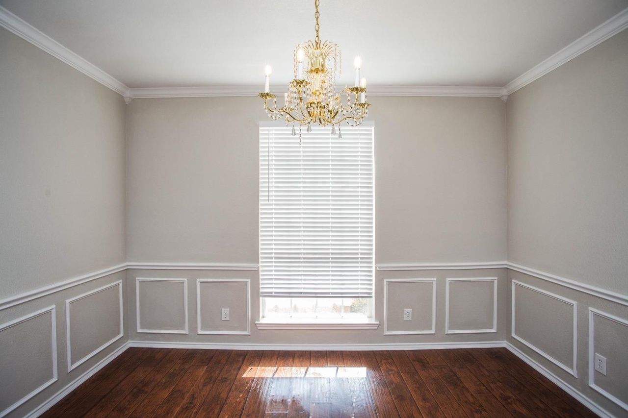 an empty dining room with a golden chandelier hanging from the ceiling. the room features white wall paneling and crown molding, and hardwood floors. there is a window with blinds in the center of the back wall, and electrical outlets are visible on the walls. the walls are painted a light gray color.