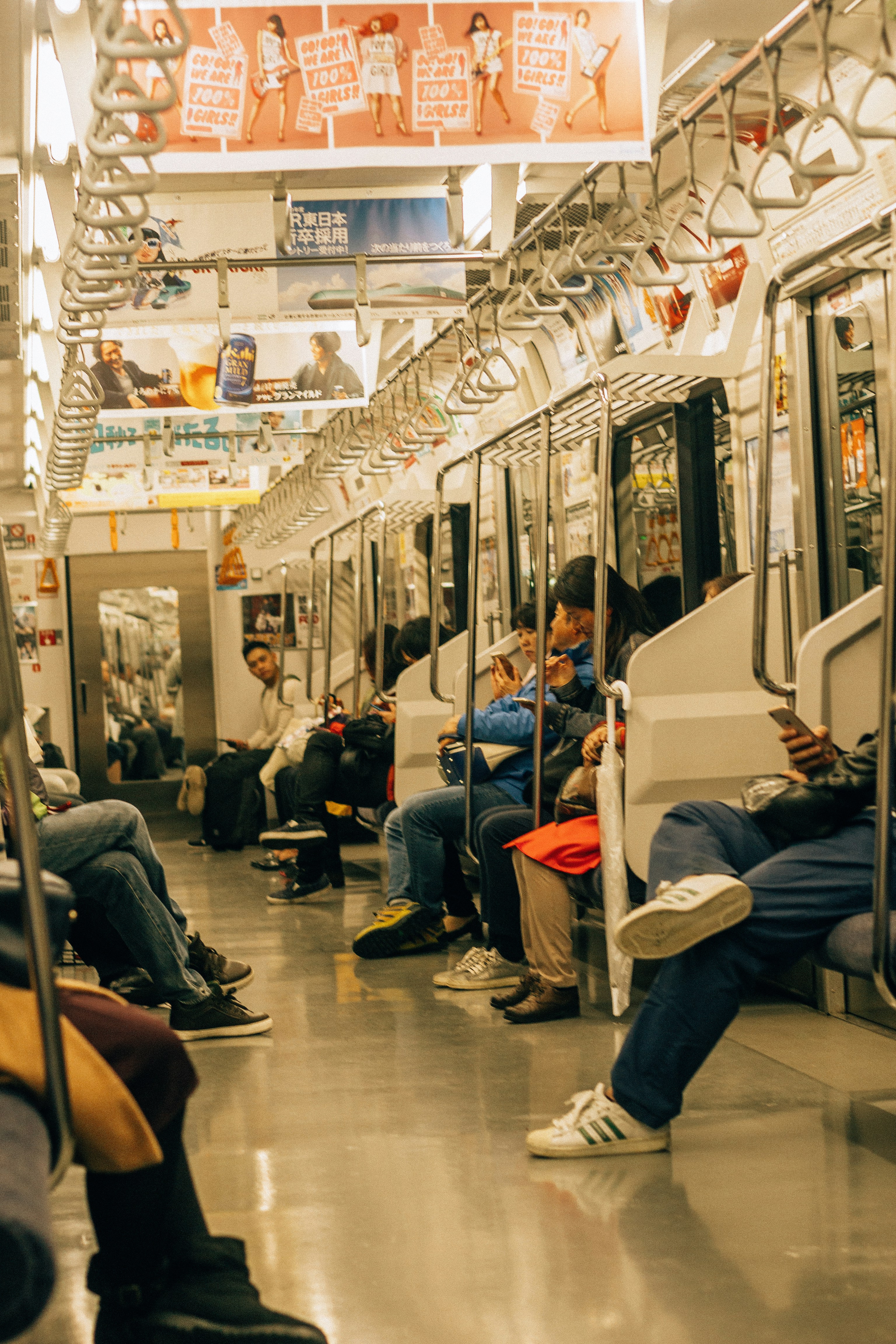 the image shows the interior of a train car, with several people seated. many of the passengers are using their smartphones. handrails hang from the ceiling for standing passengers. advertisements are visible along the top of the train car. the floor is a shiny, reflective surface. the overall lighting appears warm.