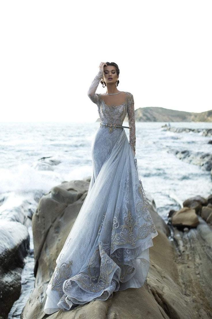 a woman wearing a beautiful silver and blue gown poses on a rocky beach near the ocean. the dress is long and flowing with intricate lace and embroidery detailing. the model is posing with her hand on her head, looking down at the ground, with a calm expression. the background is blurred, creating a soft focus. the woman is elegant and beautiful, and the overall image is romantic and dreamy.