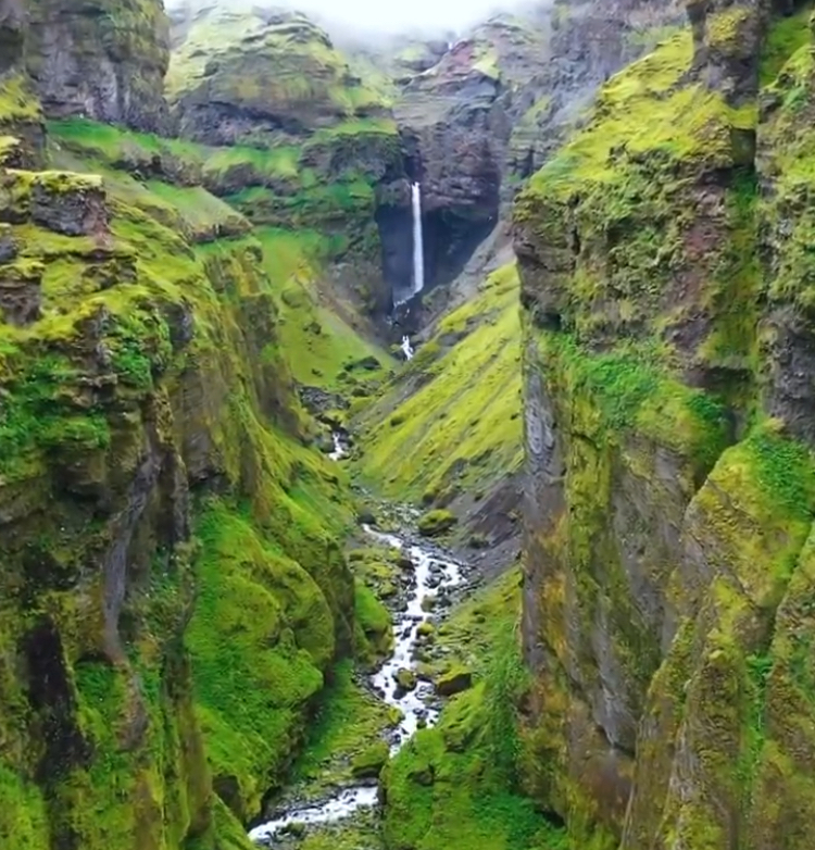 the image shows a deep canyon with a waterfall at the end. a river flows through the bottom of the canyon, and the canyon walls are covered in moss. the cliffs are tall and steep, and the overall landscape is lush and green.