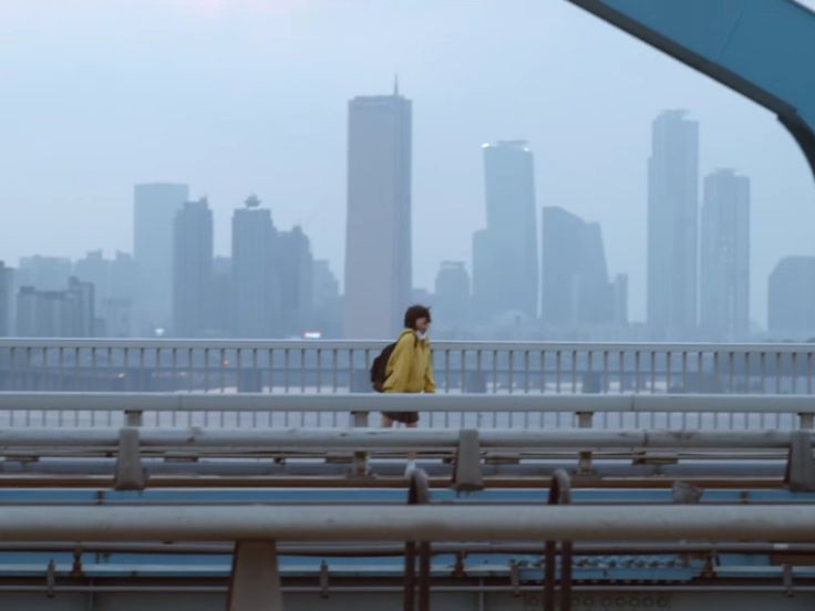 a young woman with a backpack walks across a bridge overlooking the cityscape of seoul, south korea on a foggy day.  the city's skyscrapers are visible in the background, blurred by the atmospheric haze. the woman is wearing a yellow jacket, and the overall mood is calm and peaceful. the focus is on the woman and the bridge, with the city providing a hazy backdrop.