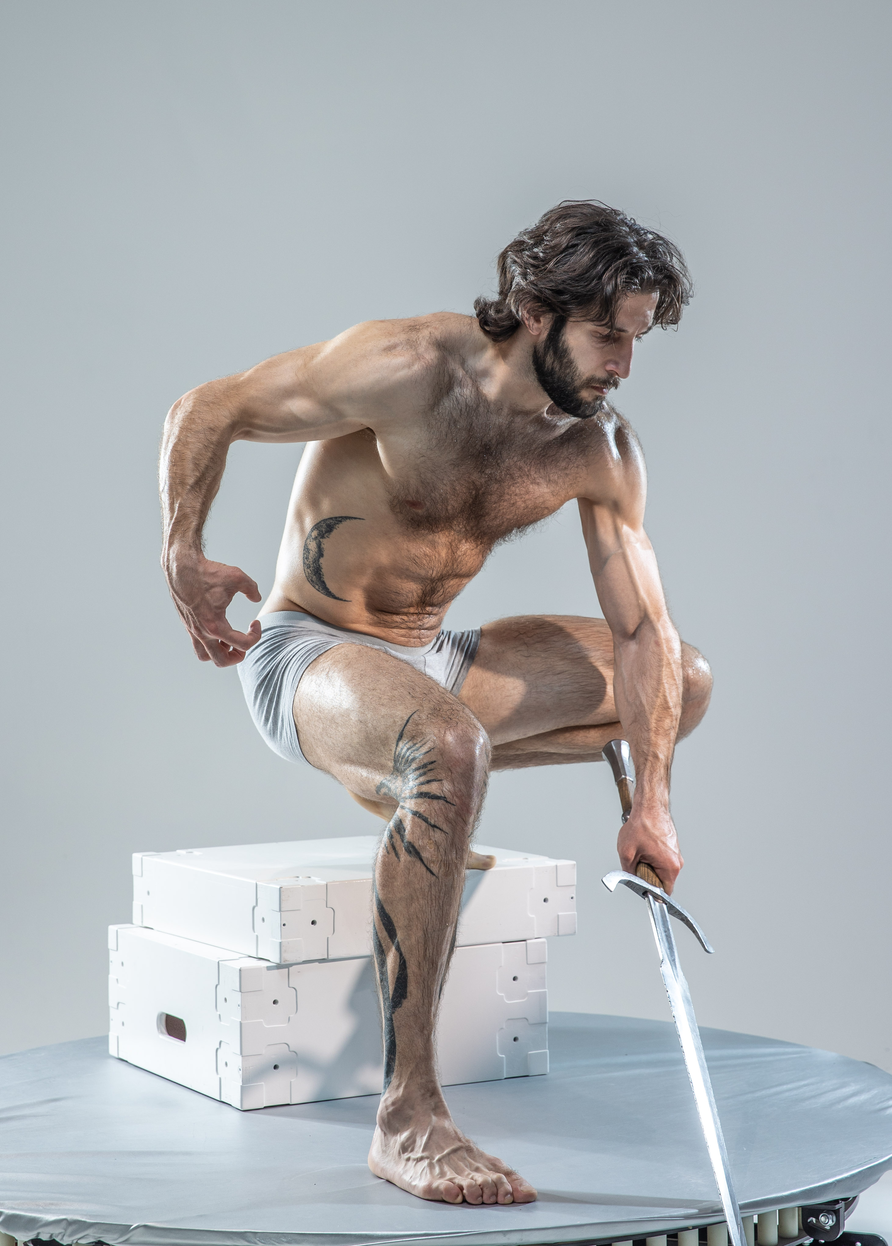 a muscular man with long hair and beard is captured midpose in a studio setting. he is shirtless, wearing only white underwear, showcasing his physique and tattoos. he holds a long sword in his right hand, seemingly about to strike, while his left hand rests on his thigh. he crouches on white boxes, positioned on a large round platform. the image emphasizes strength and action, with studio lighting highlighting the model's muscles and the sword's metallic sheen.