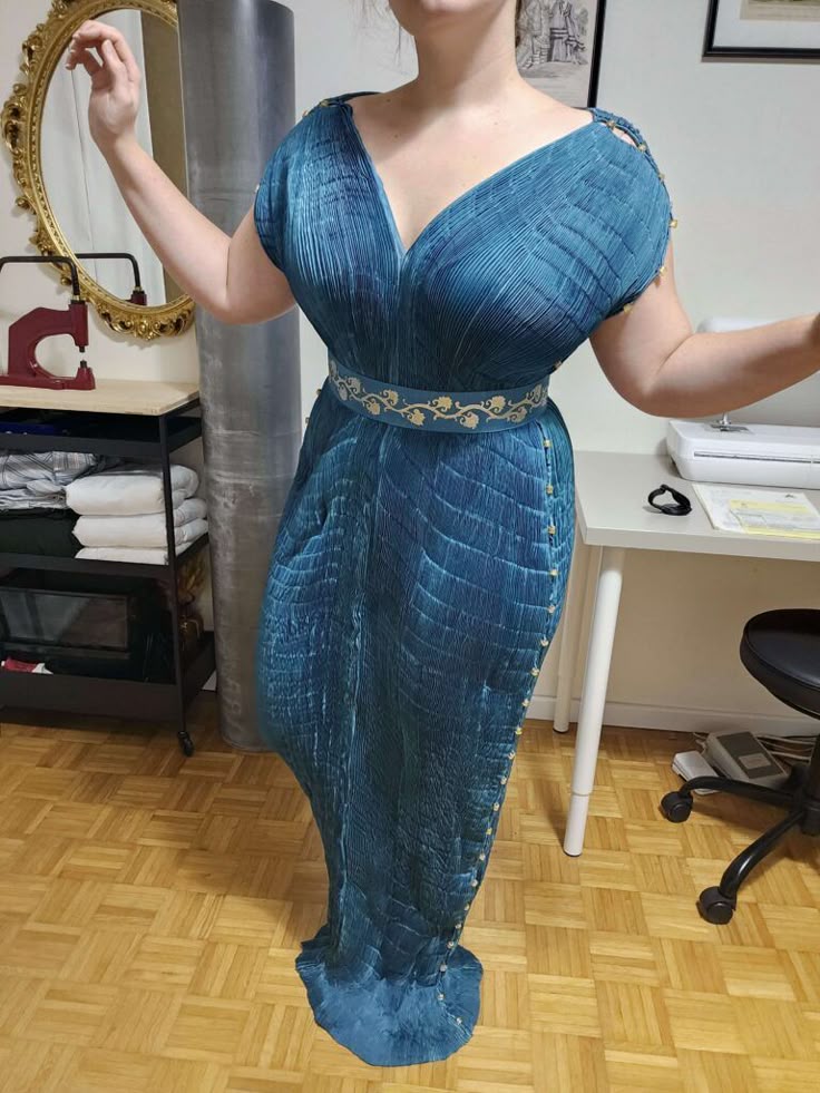 a woman is wearing a long, blue, pleated dress with a vneck and a gold belt. she is standing in a sewing room with a table, mirror, chair, sewing machine, and laminate flooring.