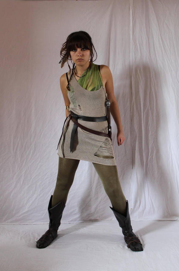 a fullbody shot of a young woman dressed in a fantasystyle costume. she is wearing a beige tunic with rips and holes, olive green leggings, tall dark boots, and a brown leather belt with a sword or dagger strapped to her hip.  her hair is dark brown with bangs framing her face. she has a serious expression. the background is a plain white fabric backdrop.  the overall style suggests a medieval or postapocalyptic warrior or adventurer character.