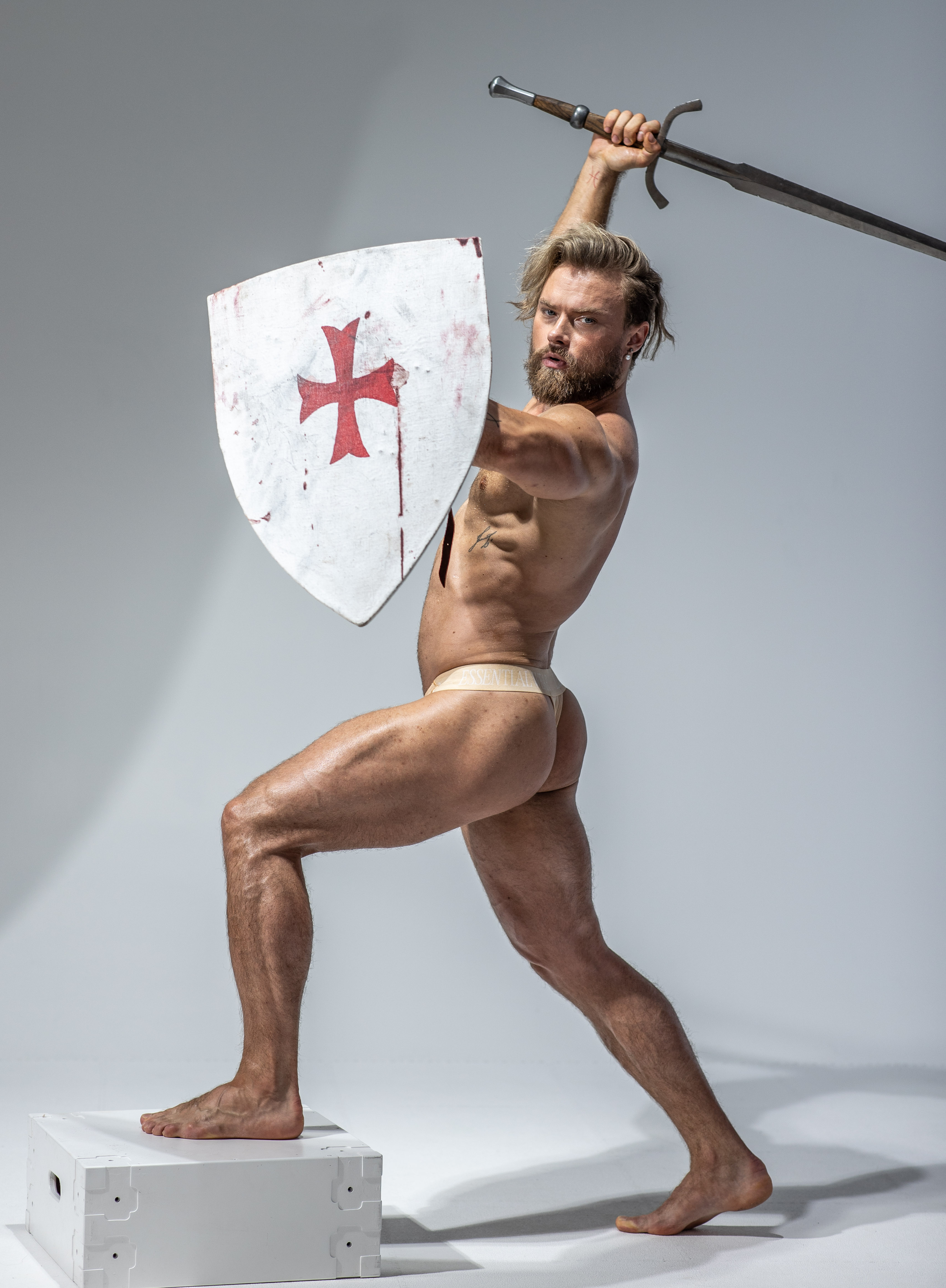 a muscular man stands in a dynamic pose on a white platform, wearing only beige underwear. he holds a sword raised in his right hand, ready to strike, and a shield with a red cross in his left hand, suggesting a medieval knight or warrior. the scene is set in a studio with soft lighting and a plain white background, highlighting the man's physique and the details of his weaponry.