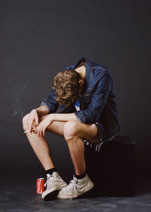 a young man sits on a black box with his head down and arms resting on his legs, looking sad and contemplative. he is wearing a denim jacket, jean shorts, and white sneakers.  the image is simple and minimal, with a black background.