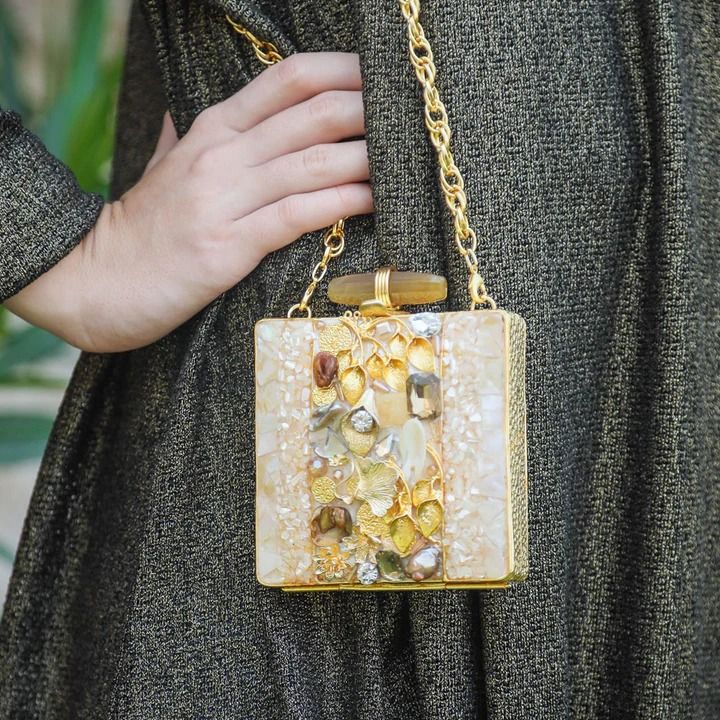 a woman is holding a square handbag with a gold chain. the bag is decorated with beads, rhinestones, and a horizontal wooden bar on top. the woman is wearing a dress made of gold fabric, and her hand is resting on the bag.