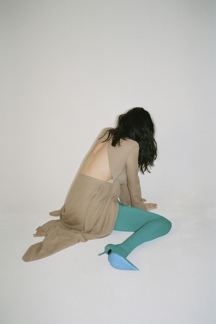 a woman is sitting on the ground, wearing a brown dress with long sleeves, and teal blue tights with matching heeled shoes. she is sitting on a white background, and her face is out of view.