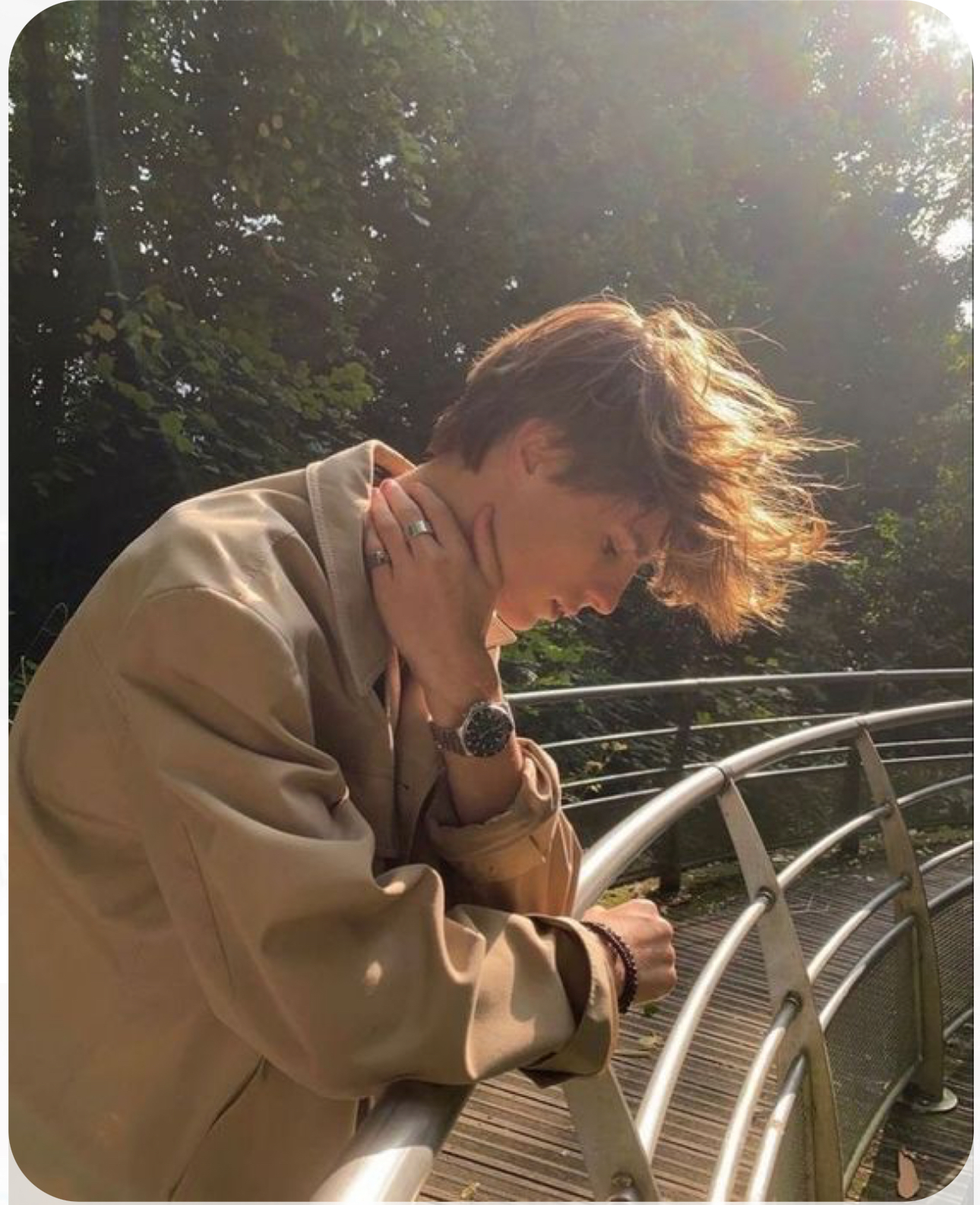 a young man wearing a light brown jacket with his hand on his neck is leaning on the railing of a bridge. he's looking down and his windblown hair is partially obscuring his face.  the sunlight shining through the trees behind him gives the image a warm, golden glow.