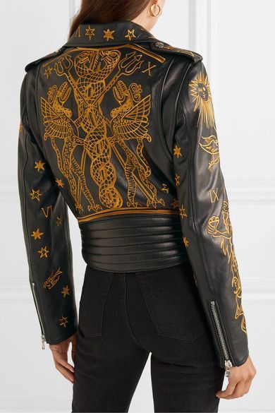 a black leather jacket with goldcolored print worn by a woman. the print features stylized depictions of snakes, eagles, and other symbols. the jacket has a zipper closure and detailing on the sleeves and back.