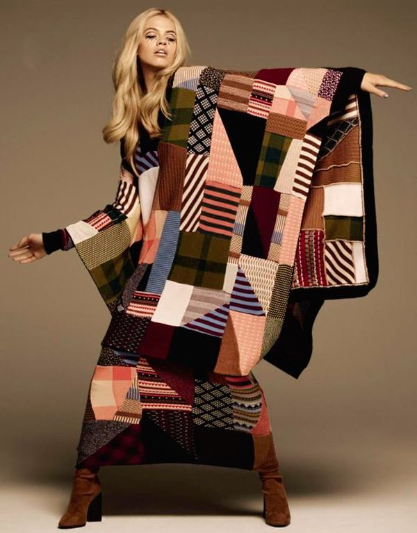 a blonde woman in a long, patchwork quiltstyle dress with long sleeves and a matching skirt poses in a studio with a beige background. she is wearing kneehigh brown boots and has a neutral makeup look with her hair styled in loose waves.