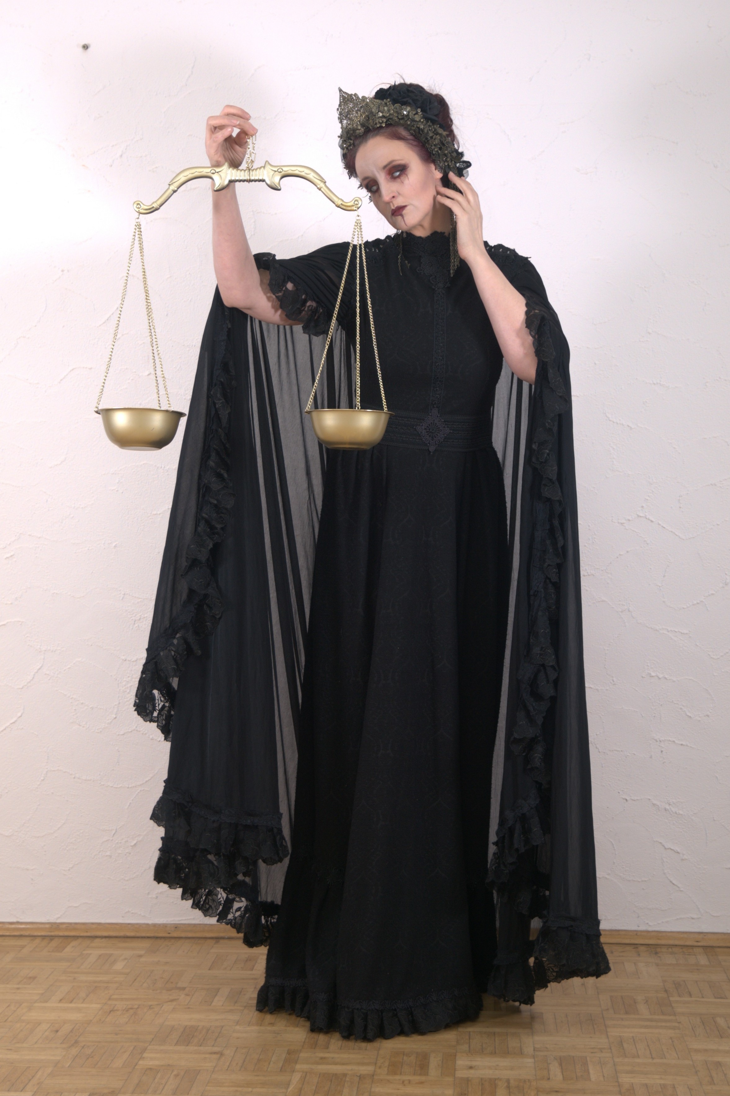 a woman dressed in a long black gown and a crown stands holding scales in her hands. she is in costume, reminiscent of a personification of justice or fate. her gown features ruffles along the sleeves and hem. the gold scales hang from a matching golden rod. the woman has pale skin and dark makeup. she stands on a wood floor against a plain offwhite wall.
