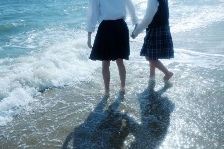 the image shows two schoolgirls, likely japanese, wearing their school uniforms, walking handinhand along a beach where gentle waves are lapping the shore. they are barefoot and their shadows are visible on the wet sand. the overall mood is serene and peaceful, evoking a sense of friendship and carefree summer joy. the focus is on the girls' back, only showing legs and waists. the background is blurred but clearly shows a light blue ocean and foaming waves.