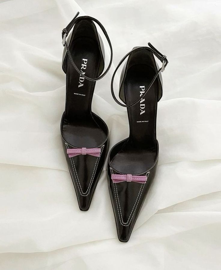 a pair of black leather prada heels with pointy toes and ankle straps. the heels have a small pink bow on the top of each shoe. the shoes are resting on a white surface.