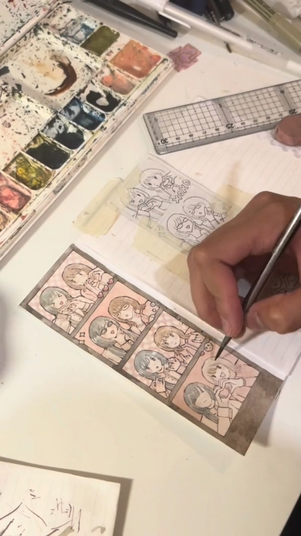 the image shows an artist drawing manga illustrations of a group of girls in a photo booth strip. the drawing is done on a table with various art supplies surrounding it.