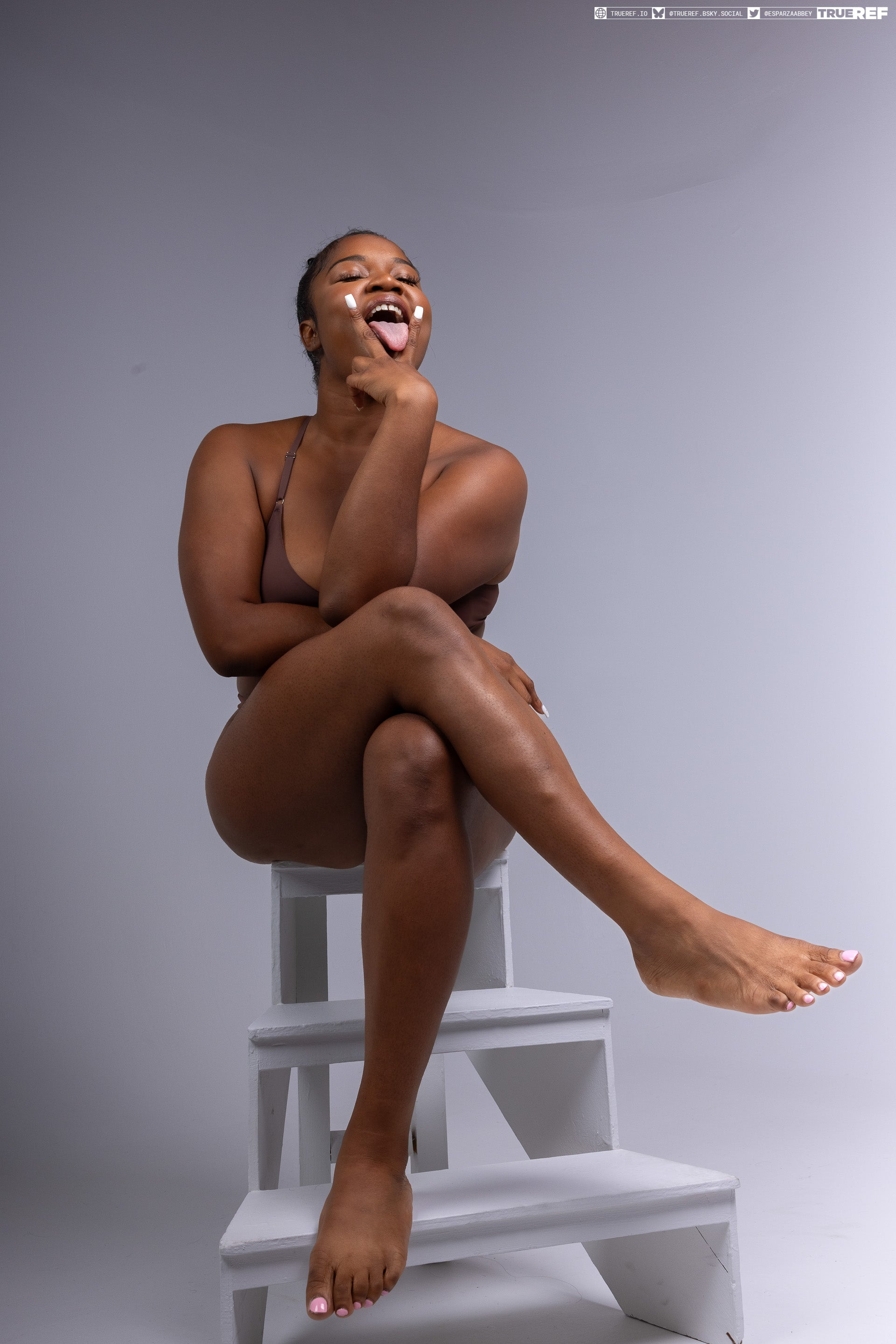 a darkskinned woman in a brown bra is sitting on steps, full shot. her legs are crossed, and she's sticking out her tongue with white facial cream on her face. the background is a plain gray studio. she has her hand near her face. she is looking up and appears playful.