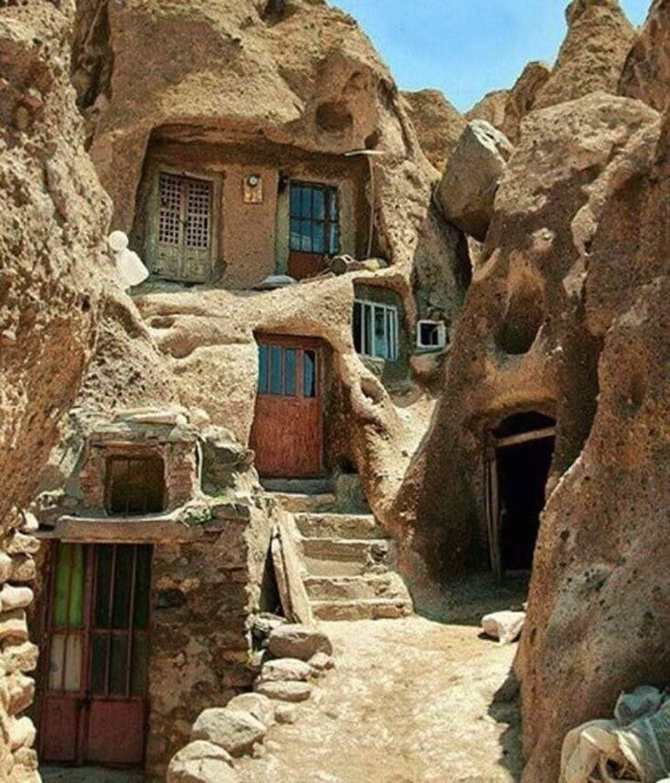 the image shows the kandovan village, a historical troglodyte village in iran known for its cave dwellings carved into rock formations. the structures appear as multilevel houses with doorways and windows, built into the soft volcanic rock of the landscape. a stone path leads through the village, with steps connecting the different levels and homes.