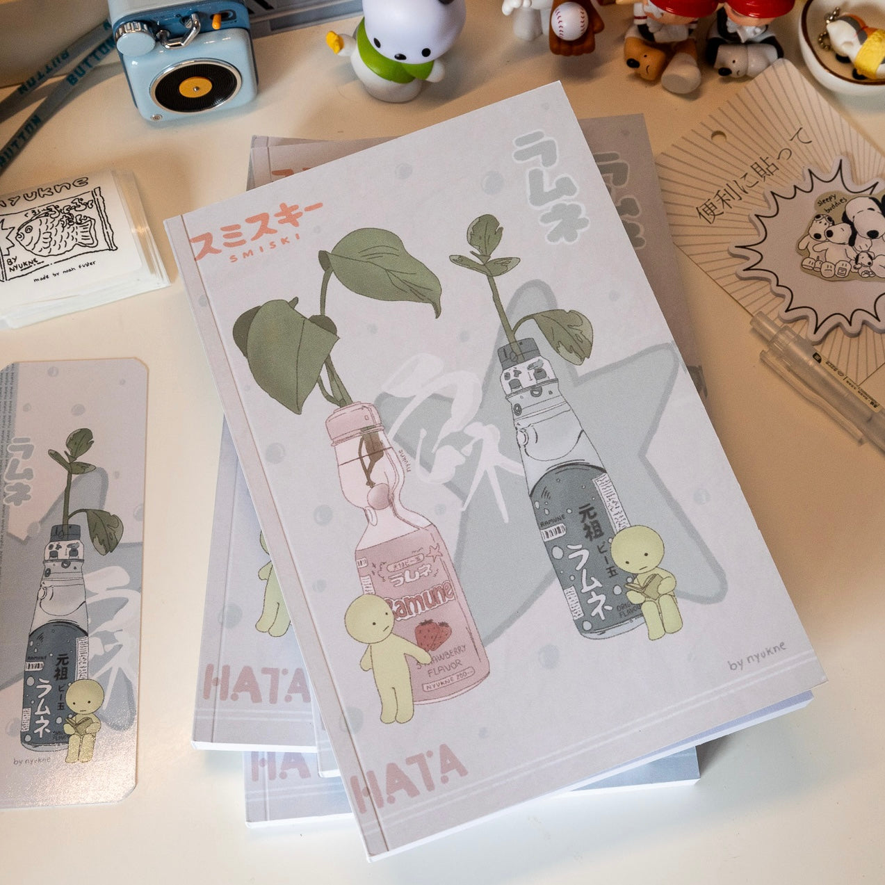 a closeup shot of a stack of notebooks featuring illustrations of ramune bottles with plants growing out of them. the illustrations are in a pastel color scheme and have a minimalist aesthetic.  the notebooks are labeled with the artist's name and the word "hata".  other stationery items and small toys are visible in the background.  a bookmark featuring a similar design is also present. the overall style is reminiscent of japanese kawaii aesthetics.