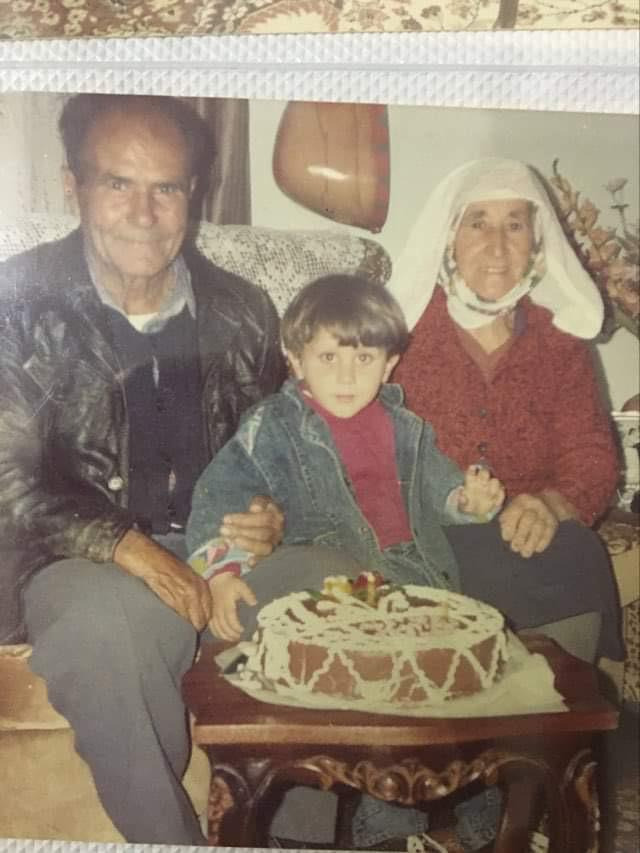 the photo shows three generations of a family gathered together.  an elderly couple, a man and a woman, are seated on a sofa with their young grandchild. the man is wearing a leather jacket, and the woman is wearing a headscarf.  the child, a boy, is sitting between them. a decorated chocolate cake sits on a small table in front of them, suggesting a birthday celebration. the setting appears to be a home living room, with a visible portion of what looks like a vintage instrument (possibly a saz) hanging on the wall. the overall feel is warm and reminiscent of a cherished family memory captured in a photograph from the 1980s or 1990s.