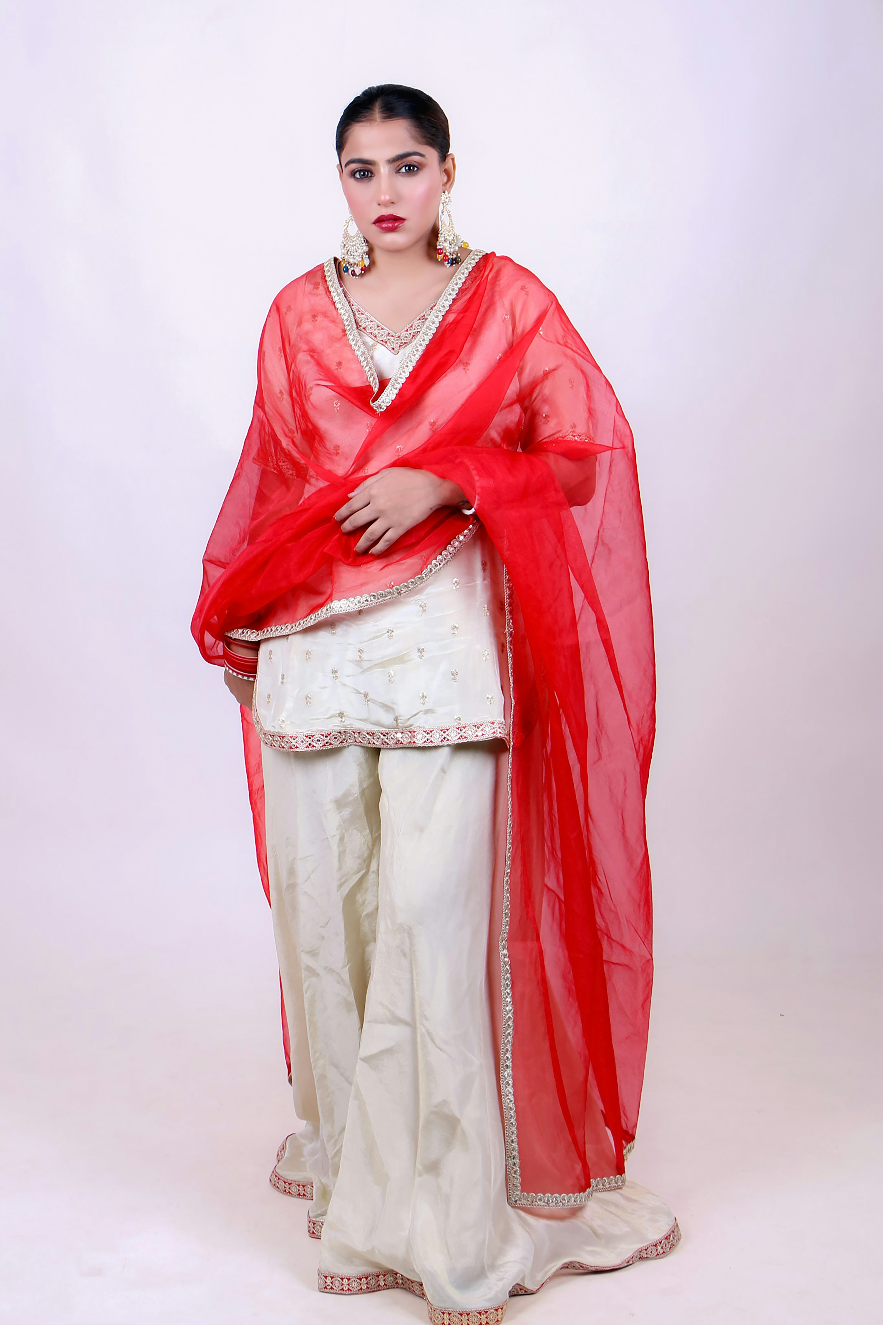 the image showcases a south asian woman modeling a stylish offwhite shalwar kameez ensemble. the kurta features delicate embroidery, paired with widelegged cream palazzo pants.  a vibrant red dupatta (shawl) with silver embellishments adds a pop of color and complements the outfit. the woman is accessorized with pearl earrings and appears to be posing for a fashion photograph in a studio setting against a white backdrop. the overall style suggests festive or traditional wear.