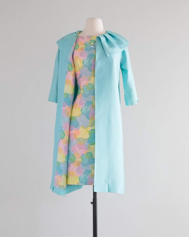 a vintage floral dress is displayed on a mannequin, along with a light blue coat. the mannequin is also wearing a necklace.