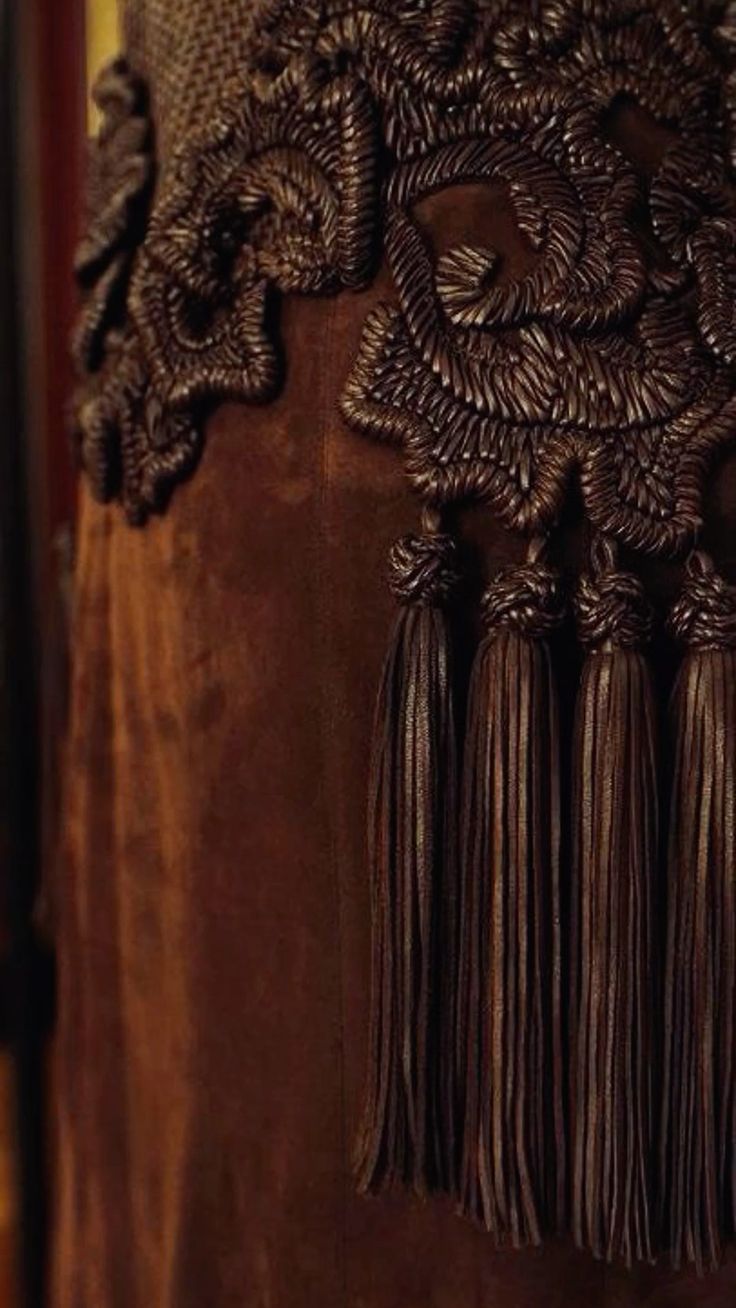 this is a closeup image of a brown leather fringe, tassel and suede fabric. the fringe is intricately woven with brown leather cord. the tassels are also made of leather and hang from the fringe, creating a luxurious and elegant look. the suede fabric is soft and rich, adding to the overall luxurious feel of the image. the image is a beautiful example of handmade craftsmanship and detail.