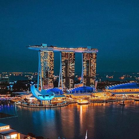 the image showcases a stunning night view of the marina bay sands integrated resort in singapore.  the iconic three towers, connected by a rooftop sands skypark, are brightly illuminated against the dark night sky. the surrounding waterfront area is also lit up, highlighting the modern architecture and reflecting the city lights on the calm water. the artscience museum is visible in the foreground. the overall scene is one of vibrant city life and architectural splendor.