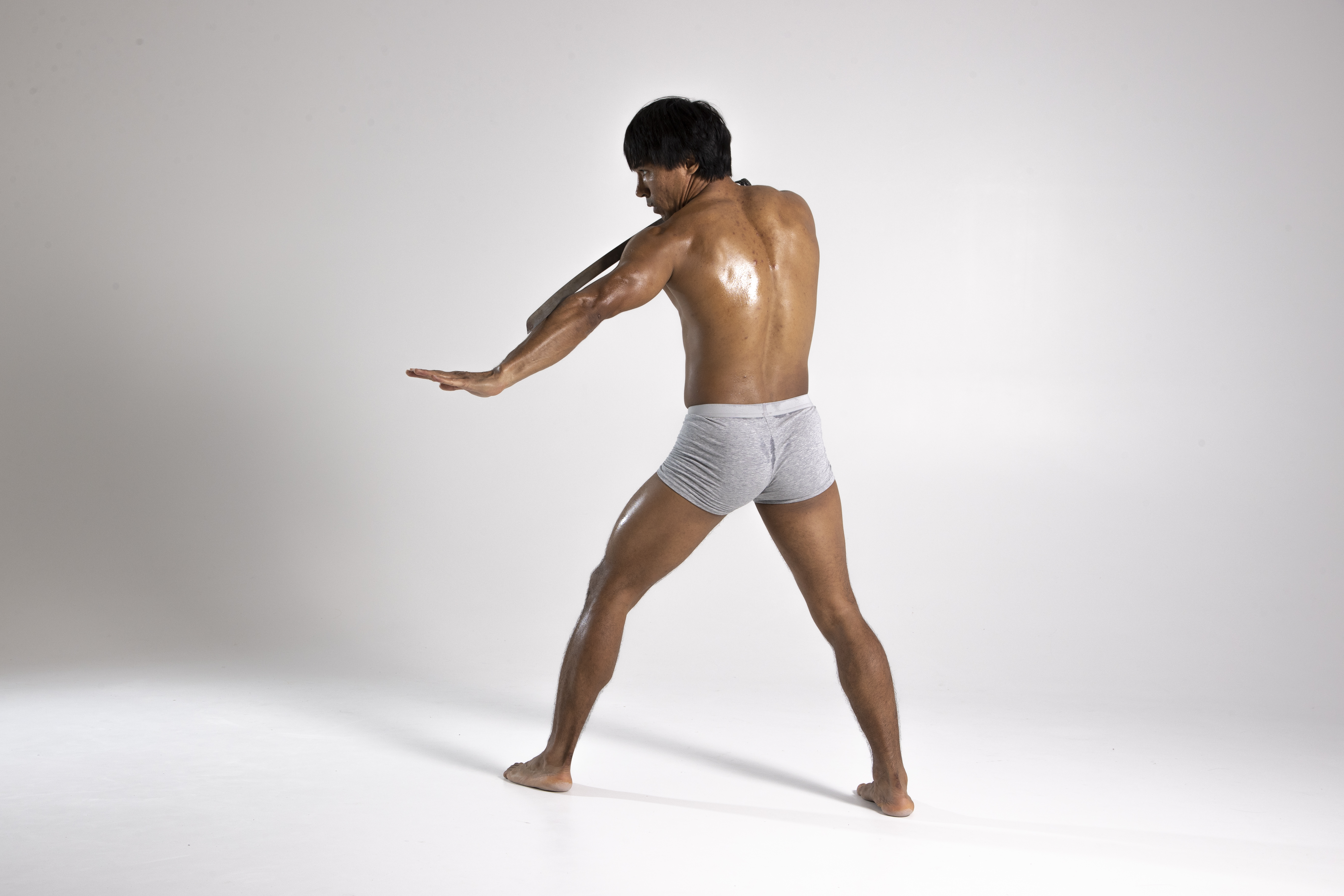 a muscular, wet man stands in an open stance with his arms extended in a studio shot against a gray background. he is wearing gray underwear and is facing away from the camera. he is asian with dark hair. his back is visible and his skin appears to be glistening. he seems to be stretching or performing a similar motion.