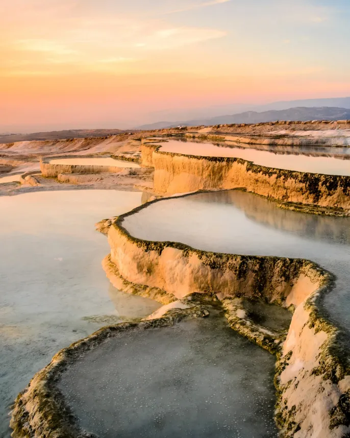 the image shows the pamukkale travertines in turkey, with its terraces of white calcium carbonate formations filled with water. the scene is bathed in the warm light of a sunset, creating a beautiful landscape.