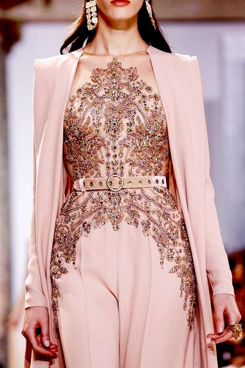 the image showcases a model on a fashion runway wearing a stunning, highfashion ensemble. the outfit is a widelegged blushpink jumpsuit with a sheer, rosegold embellished bodice. the embellishments consist of intricate beading and sequins, creating a luxurious, sparkly effect.  a matching blush pink blazer or long overcoat drapes over the jumpsuit. a thin, rose goldtoned belt cinches the waist. the model is also wearing large, dangling gold earrings. the overall aesthetic is elegant, glamorous, and sophisticated. the background suggests a highend fashion show setting.