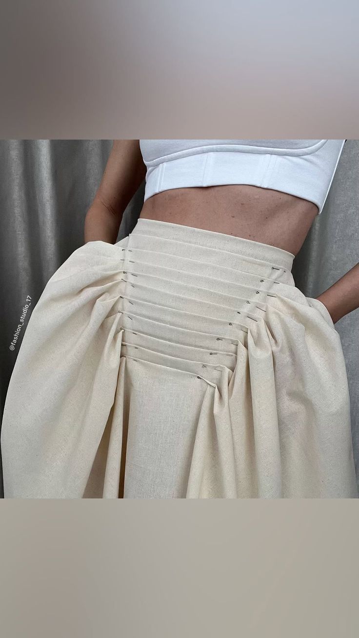 a woman is wearing a highwaisted beige skirt with intricate pleating and draping details. the skirt is paired with a white crop top. the image captures the skirt's design details, showing the construction and fabric. pins are visible along the pleats, suggesting it is a work in progress or sample garment. the backdrop is a simple studio setting.