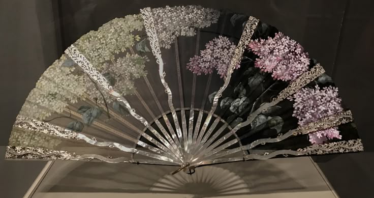 the image shows an antique hand fan decorated with a floral pattern. the fan is open, revealing a series of segments painted with different colors of lilacs that transition from white to light purple and finally to deep purple against a dark background. the edges of the fan are embellished with intricate lacelike details. the sticks of the fan, likely made of mother of pearl or a similar material, are visible and contribute to its ornate appearance. the fan casts a shadow on the surface below.