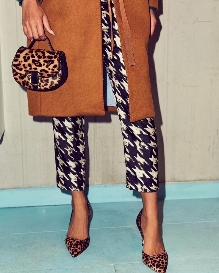 loseup view of a woman's outfit. she's wearing a camelcolored coat, black and white houndstooth cropped trousers, leopard print heels, and a small leopard print handbag.  the background is a light teal wall.  the overall style is chic and autumnal.