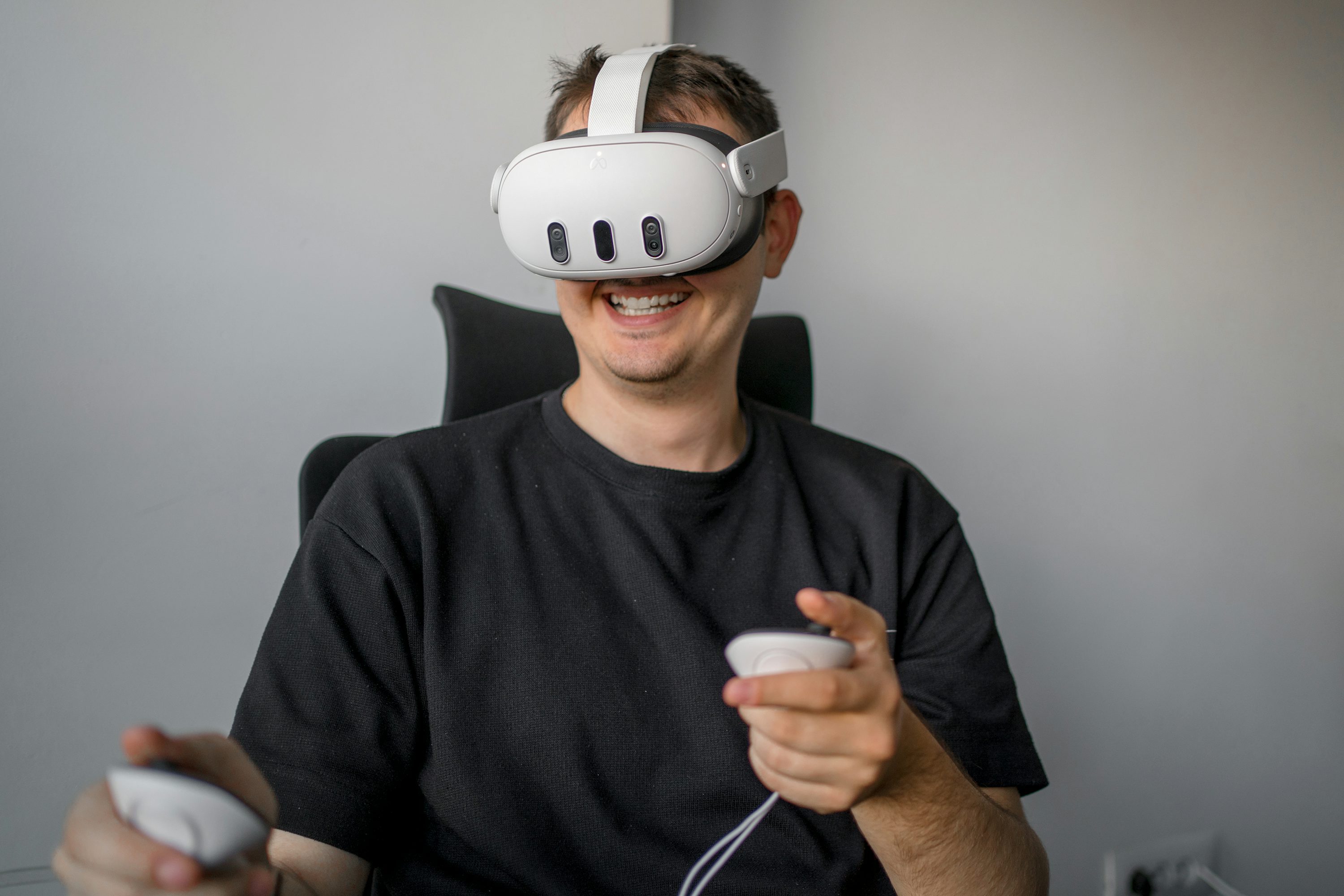 a man is sitting in a chair wearing a white virtual reality headset. he is smiling and holding two white controllers in his hands. he is wearing a black tshirt. the background is a plain gray wall.