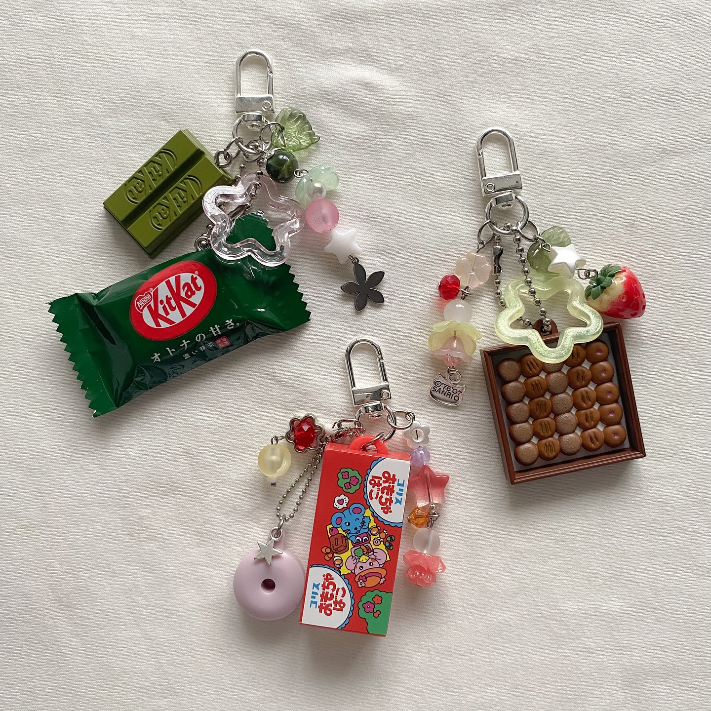 the image shows three keychains lying on a white surface. the keychains each feature miniature replicas of food items and candies. one keychain has a green tea kitkat bar and wrapper, along with green and clear beads and a flower charm. another keychain has a box of chocolate candies and a strawberry charm, also with beads. the third keychain features a red box of japanese candy with cartoon characters, a pink donut charm, and additional beads. all three keychains have silvertoned clasps.