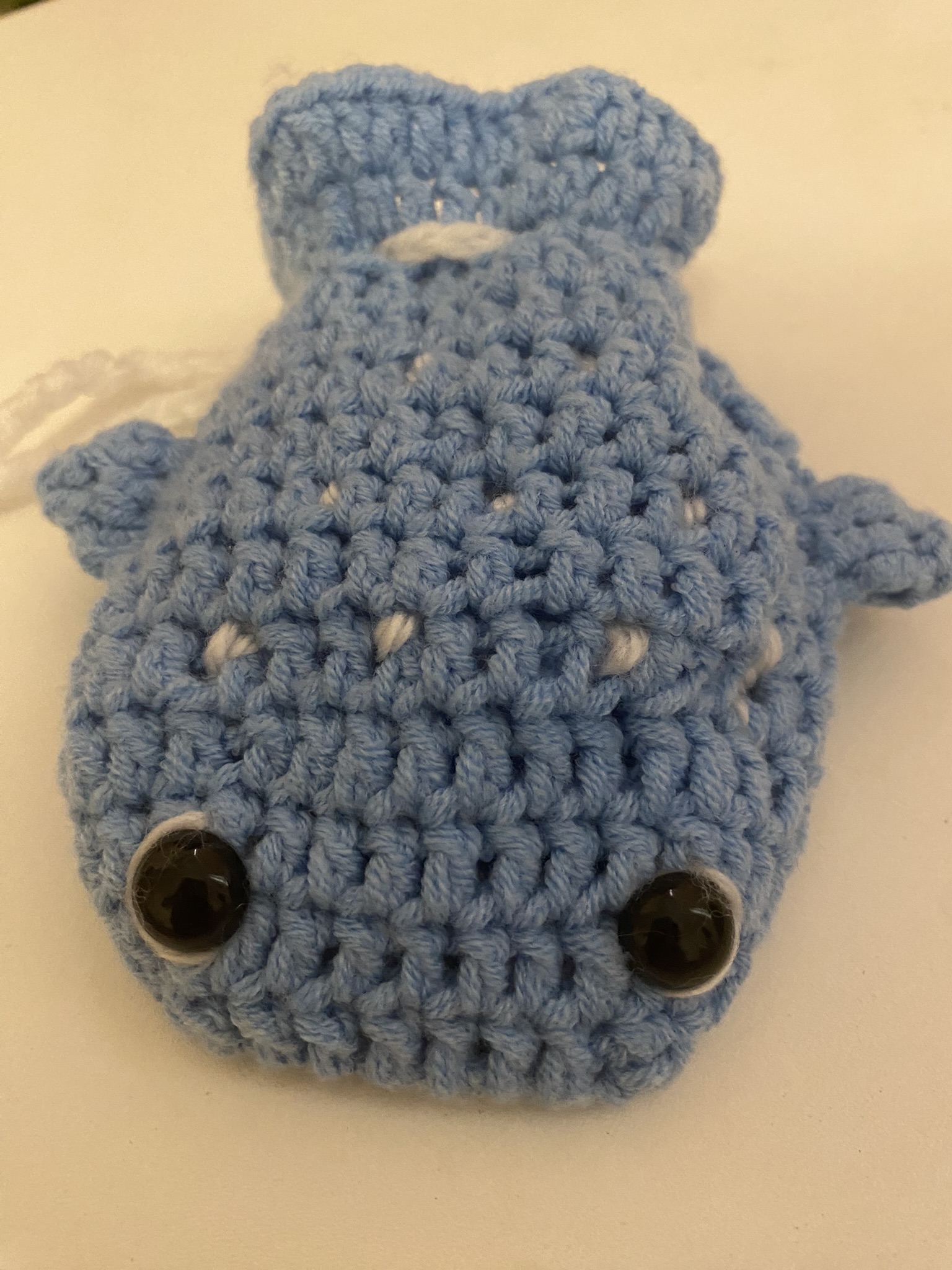 a closeup of a blue, crochet whale stuffed animal. it has large, black, plastic eyes and small white spots scattered throughout its body. the whale has small fins and a tail. a white yarn string is attached to the side.