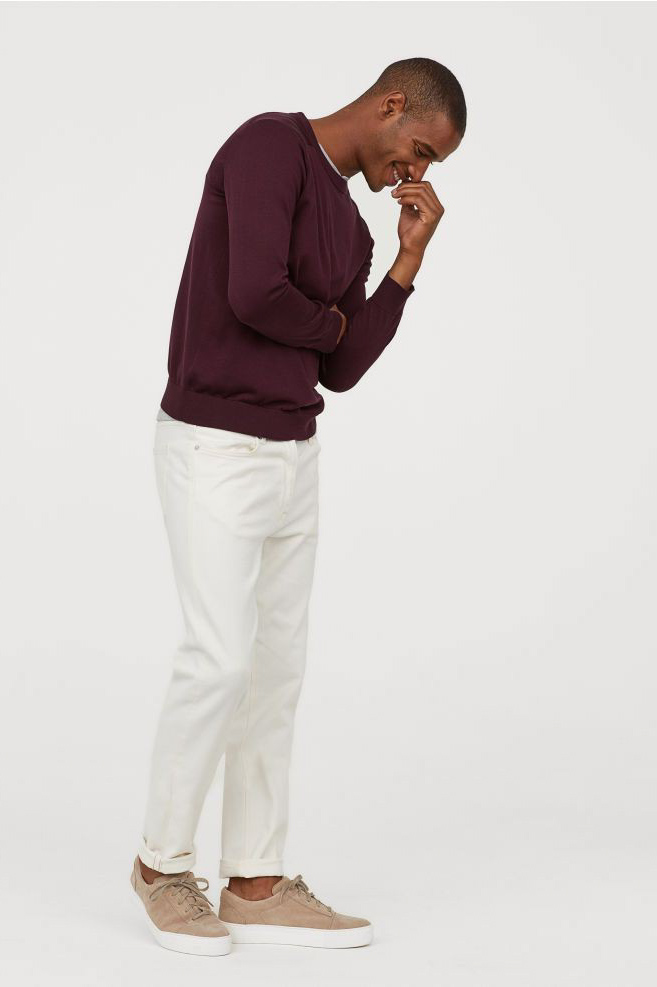 a man is wearing a burgundy sweater and white jeans. he's standing in a casual pose and smiling while looking down. he is wearing brown sneakers.