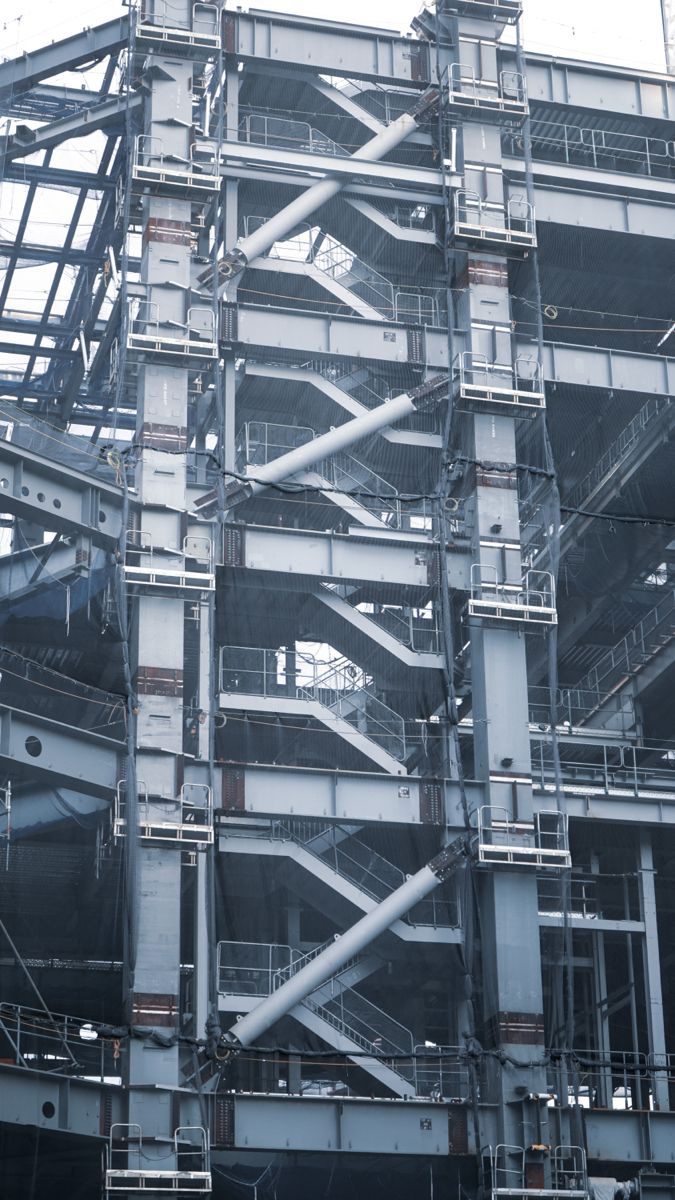 the image shows a detailed closeup view of a large steel structure, possibly part of a building under construction. the structure is composed of numerous interconnected metal beams, supports, and staircases, creating a complex and intricate network of geometric shapes.  the overall color palette is predominantly grey and metallic, reflecting the industrial nature of the construction.  the image captures a sense of height and scale, suggesting the immense size and complexity of the project. the photographic style is somewhat muted, with a focus on the texture and patterns within the structure.