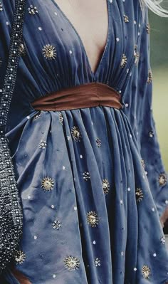 a woman is wearing a blue, embroidered,  flowing dress that has a deep v neck and a brown belt.  she is standing in the sun, and the sunlight is hitting the dress and causing it to glimmer.  the woman's face is not visible.