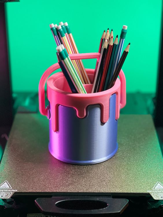a closeup of a 3d printed paint can pencil holder. the holder is pink with a blue paint drip effect. the holder is filled with a variety of colorful pencils. the 3d printer is in the background and is blurred.