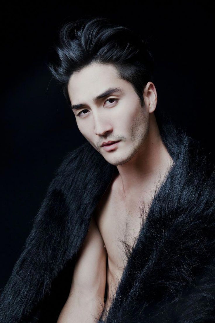 a portrait of an asian man wearing a black fur coat against a black background. he is looking at the camera with a slight smolder. he has dark hair that is styled up and back, and light stubble on his face. the fur coat is draped around his shoulders, revealing part of his bare chest.