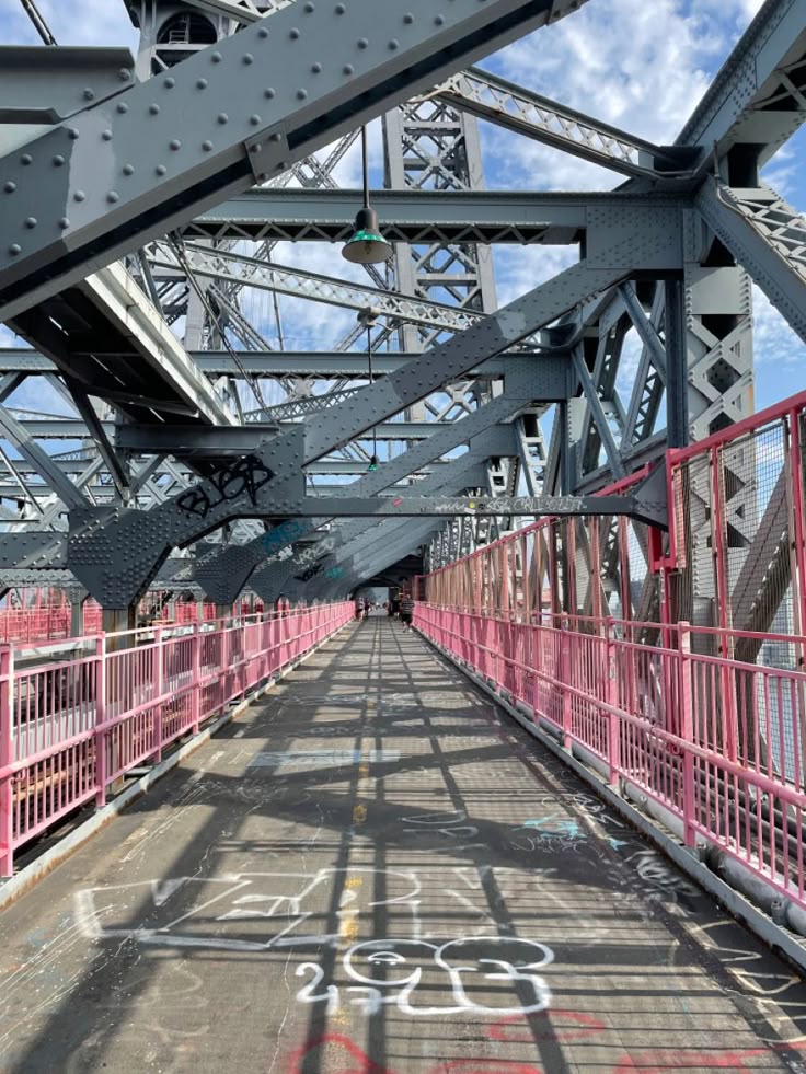 the image shows a perspective view from the middle of a bridge walkway. the walkway is made of asphalt and shows signs of graffiti. pink railings line both sides of the pathway, and the structure of the bridge itself is a prominent feature, showcasing its complex metal framework. the sky is partly visible through the steel girders. the overall setting appears to be urban, possibly in a city like new york city, given the style of the bridge. the light suggests it's daytime, with shadows cast across the walkway from the overhead structure.