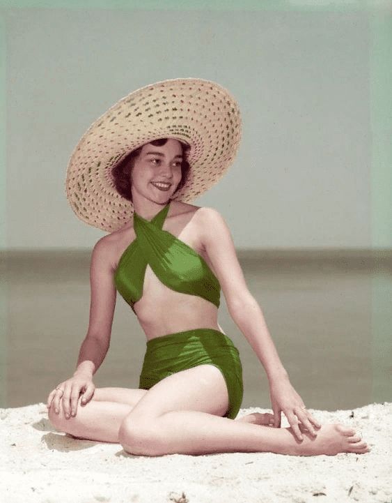 a woman in a green swimsuit and a large straw hat sits on a beach, smiling and looking to the side. the sun is shining and the sand is white. she is enjoying her time on vacation.